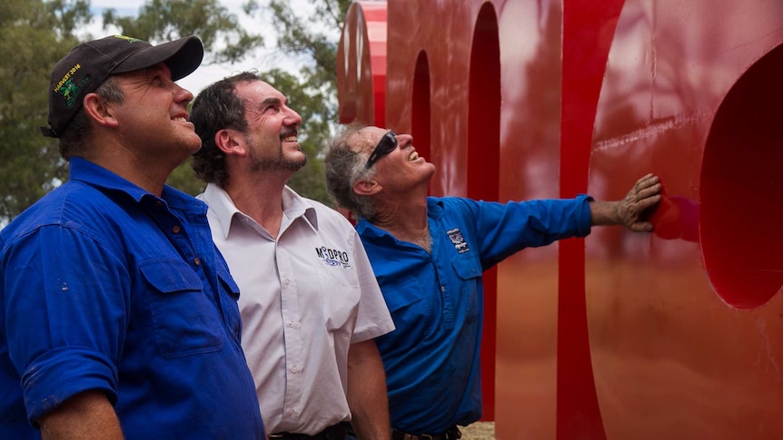 Three men looking at big shiny red letters in a bush/paddock setting