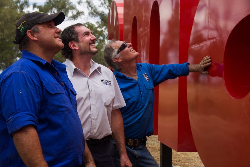 Three men looking at big shiny red letters in a bush/paddock setting