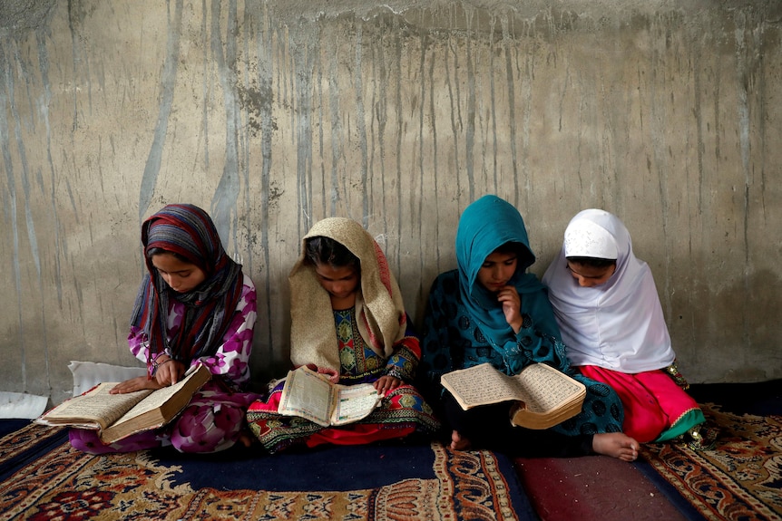 Four girls in hijabs sit on floor, heads bent, looking at Korans