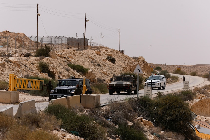 A row of military-style vehicles approach a yellow-gated checkpoint on a rocky road in bright sunlight.