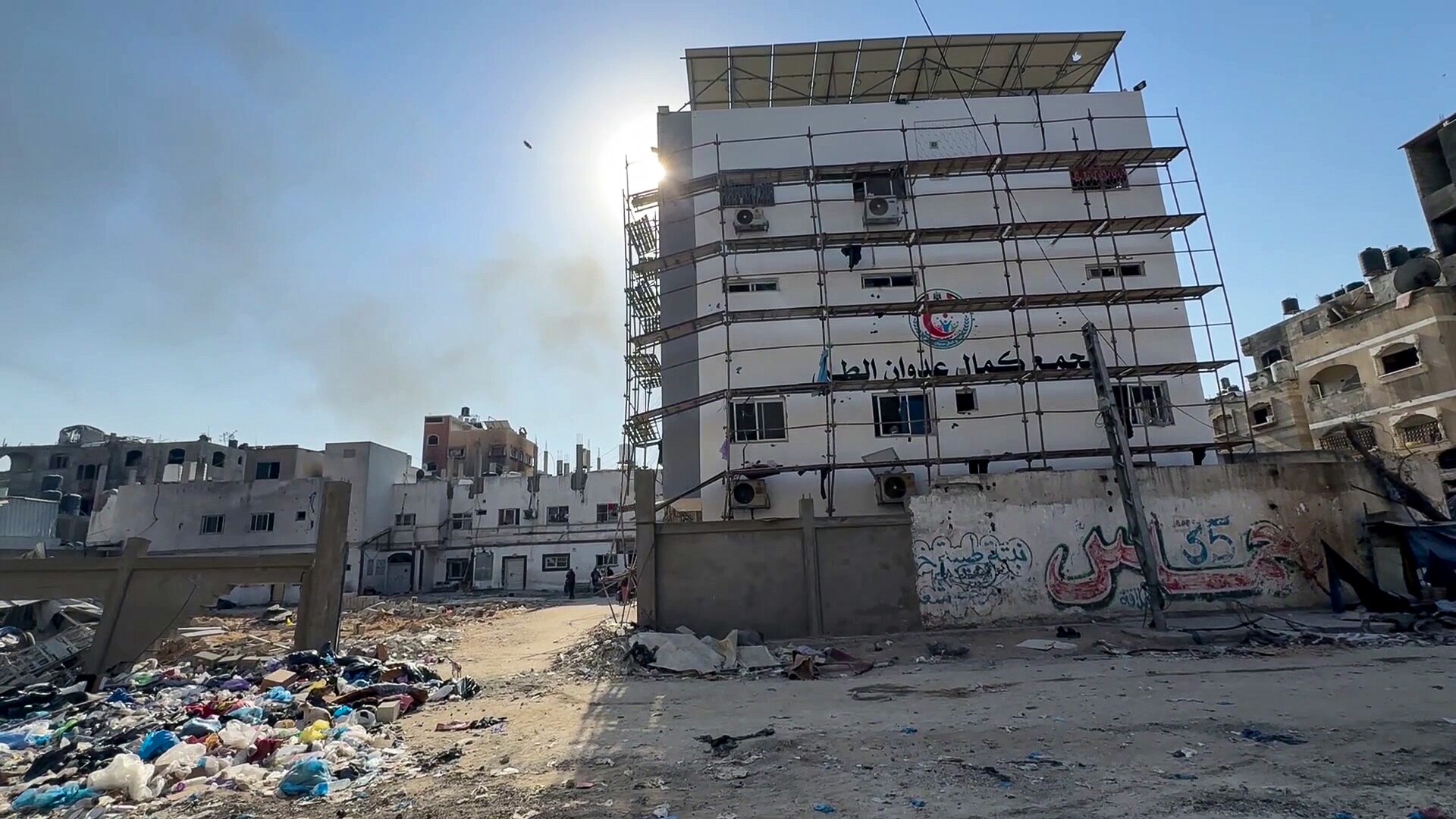 A battered multi-storey hospital with scaffolding outside sits among destroyed buildings near strewn rubbish.