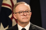 Albanese looks expectantly off camera, wearing black glasses, an Australian flag behind him