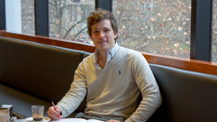 A young man sits at a cafe table with pencils and a colouring in book.