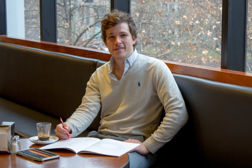 A young man sits at a cafe table with pencils and a colouring in book.