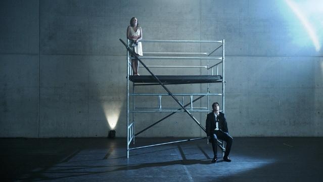Woman stands on scaffolding, man sits on scaffolding below