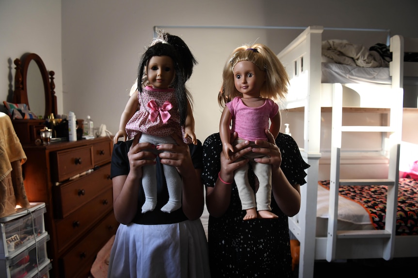 Two children hold up plastic dolls to cover their faces in a bedroom that includes bunk beds