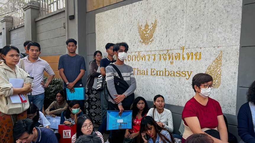 Young Asian people stand in a line outside a sign saying Thai embassy