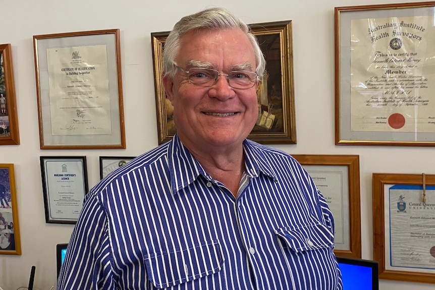 Smiling man with grey hair, wearing a blue and white striped dress shirt , standing in front of a wall full of framed documents