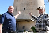 An older bald man and an older man wearing a cap stand pointing toward a large old grain silo.