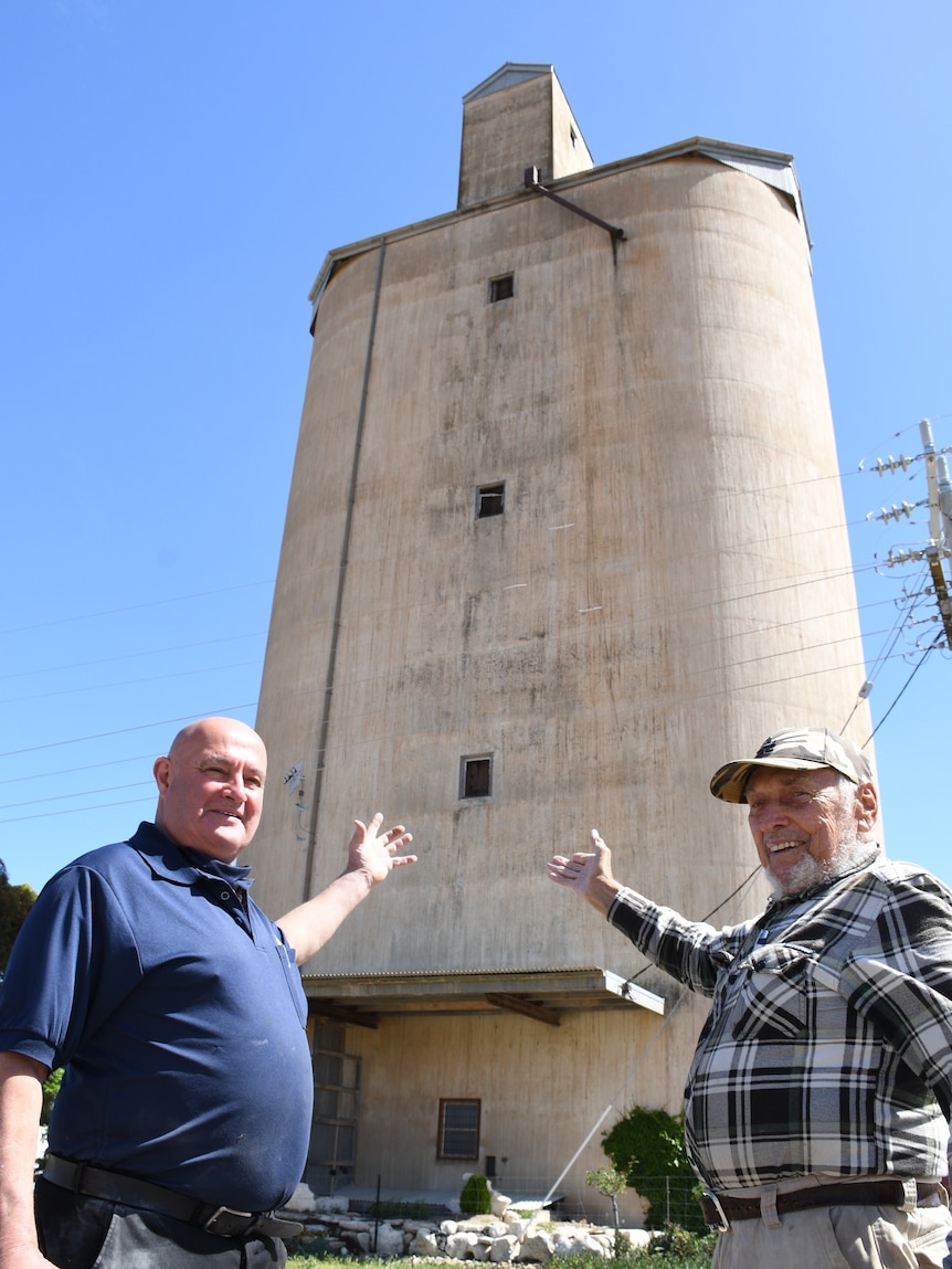 An older bald man and an older man wearing a cap stand pointing toward a large old grain silo.