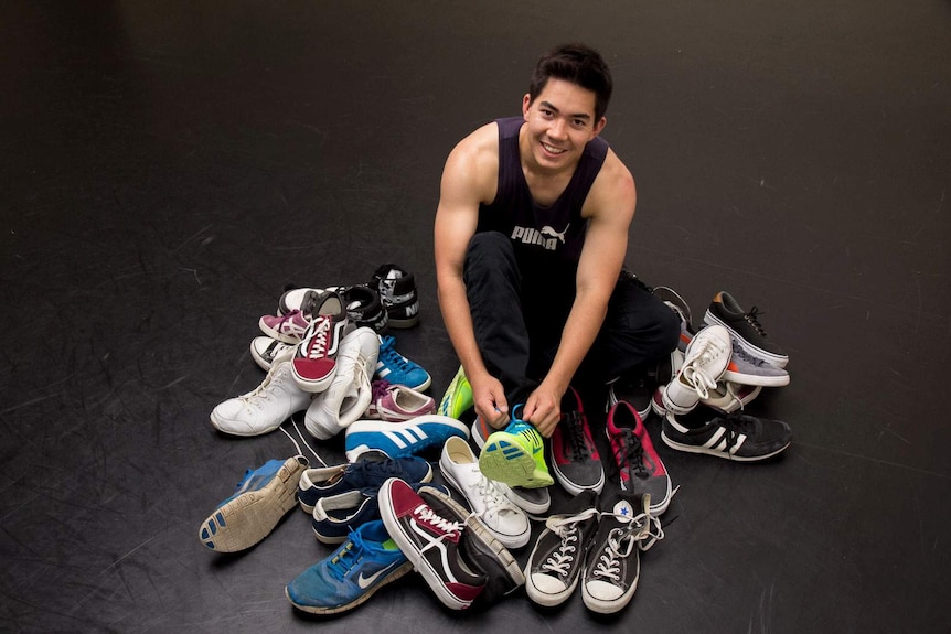 A young man in a singlet does up his sneakers surrounded by a pile of casual shoes