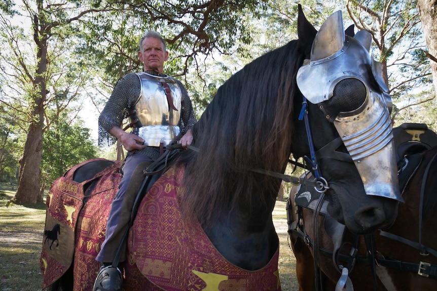 A man in shiny silver armour on a jet-black horse with trees in the background.