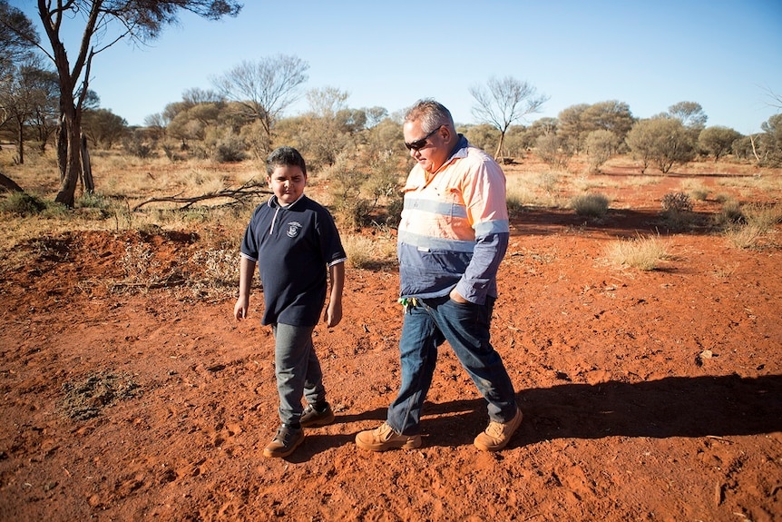 Boy walking with man across red dirt with vegetation and blue sky in background.