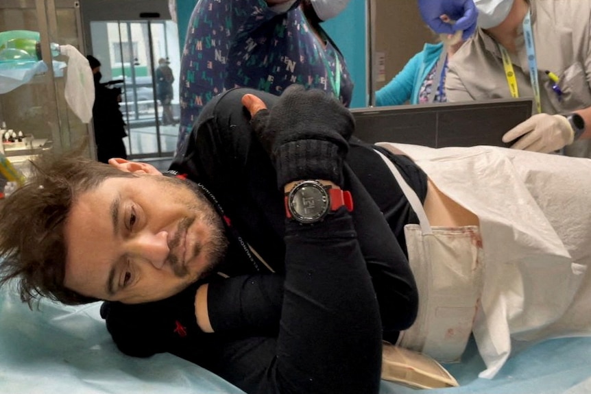 A white man lies on his side in a hospital bed, as medical personnel examine him from behind.