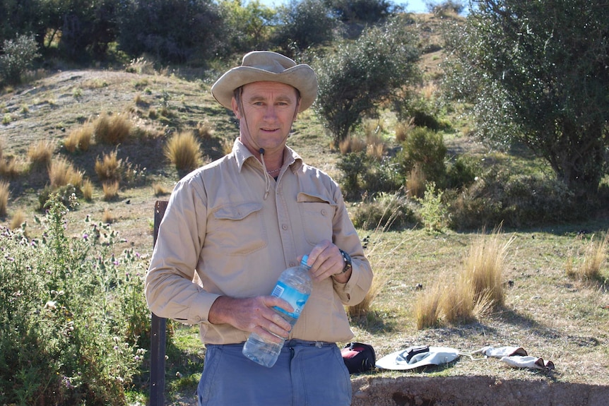 A man wearing a light brown shirt and matching hat holds a water bottle while standing in a grassy area.