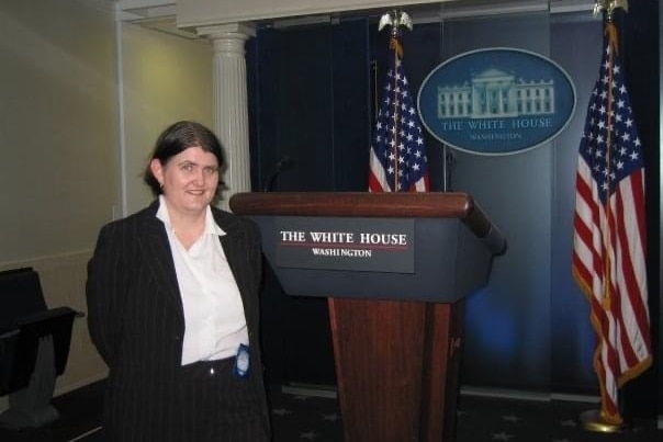 Woman posing next to White House lecturn with US flags and White House logo in background.