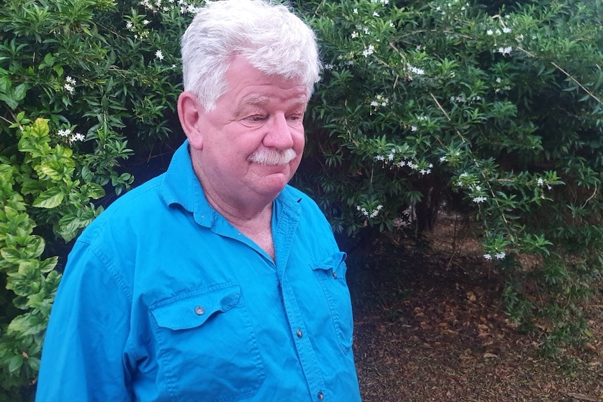 A man with grey hair and moustache in a blue collared shirt stands in front of a bush.