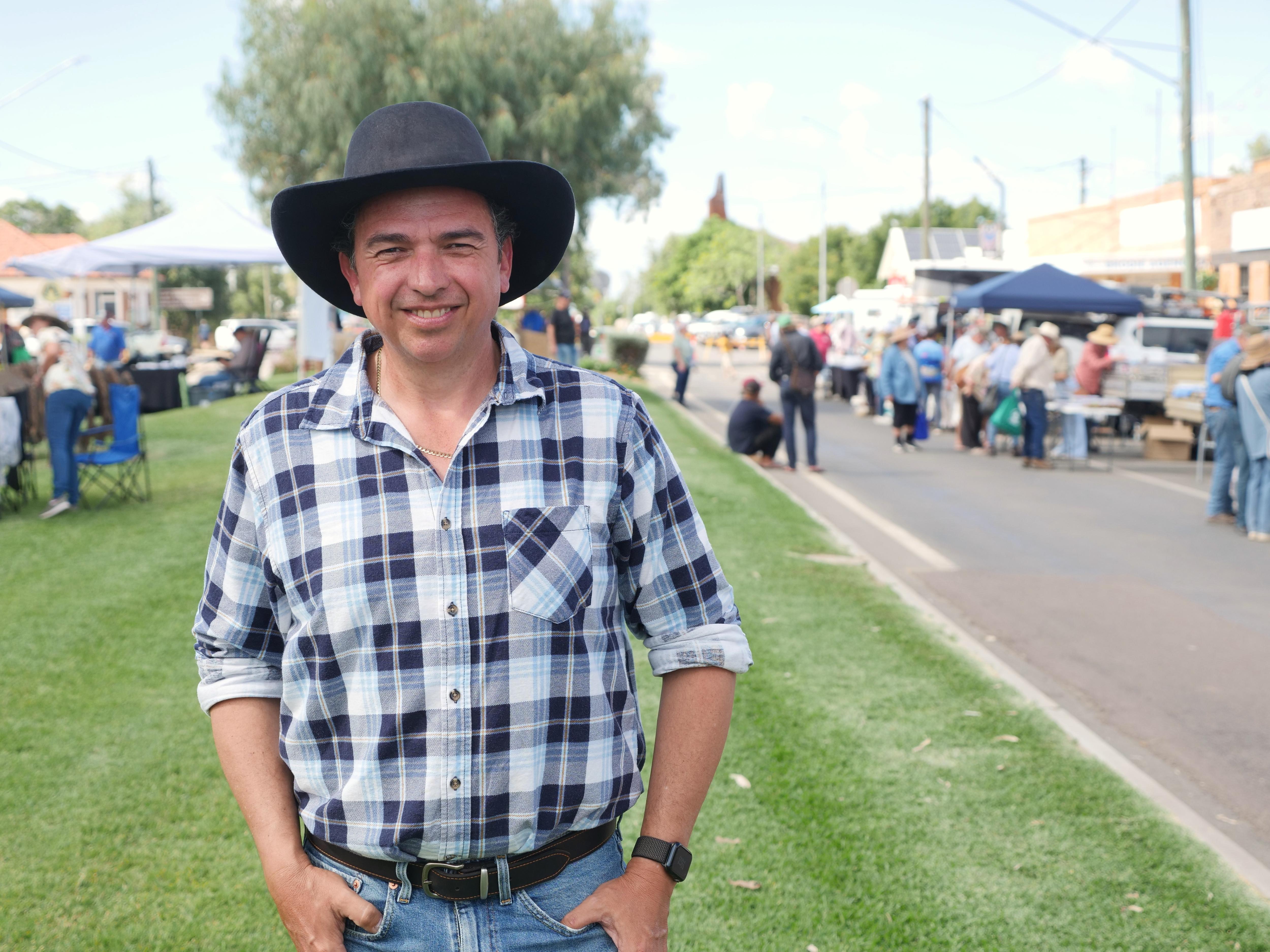A smiling man in a dark Akubra and checked shirt stands on some grass next to a street in a country town.
