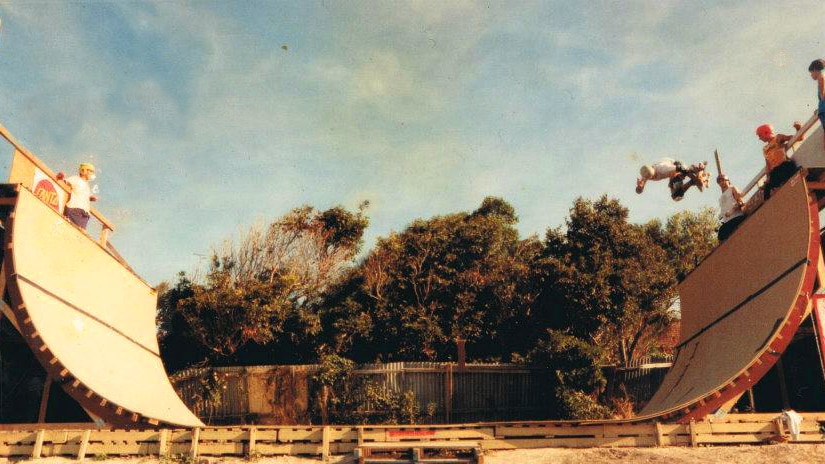 A grainy photo of a wooden skate ramp in a backyard, with a skater doing a trick in the air. 