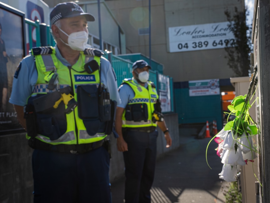 Police officers wearing a hi-vis vests and P2 masks stand in an alleyway next to a floral tribute
