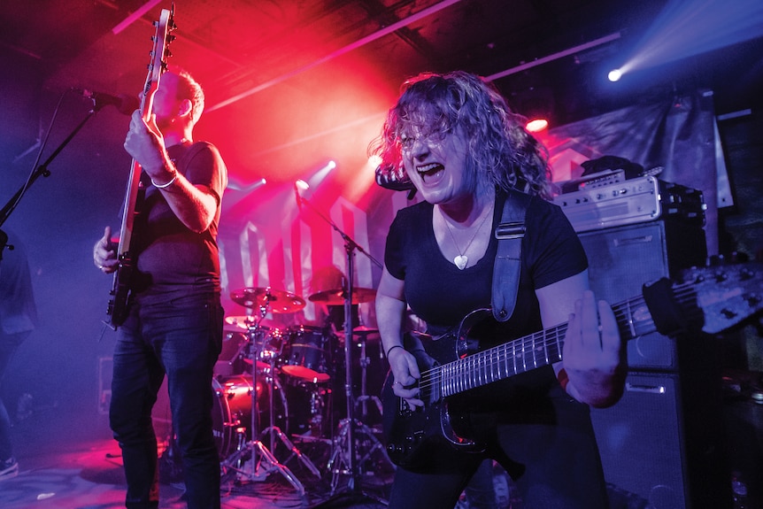 A lead guitarist sings as she plays her instrument with a bass guitarist on stage and a red light cast on the drums at the back.