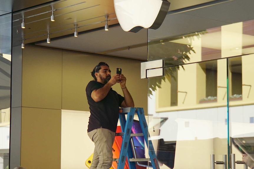 A man stands on a ladder taking a picture with a phone at the Apple Perth City store entrance with an Apple logo above him.