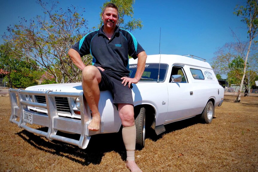A man in navy work shirt and shorts with no shoes on sits on the bonnet of a white 70s panel van larked in a backyard.