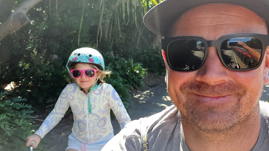 A man in sunglasses and a hat taking a selfie with a young girl riding a bicycle behind