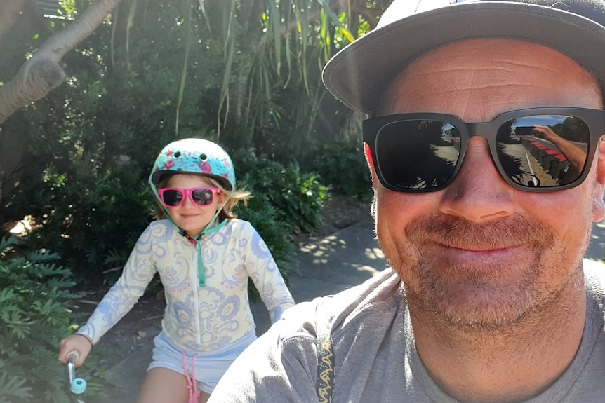 A man in sunglasses and a hat taking a selfie with a young girl riding a bicycle behind
