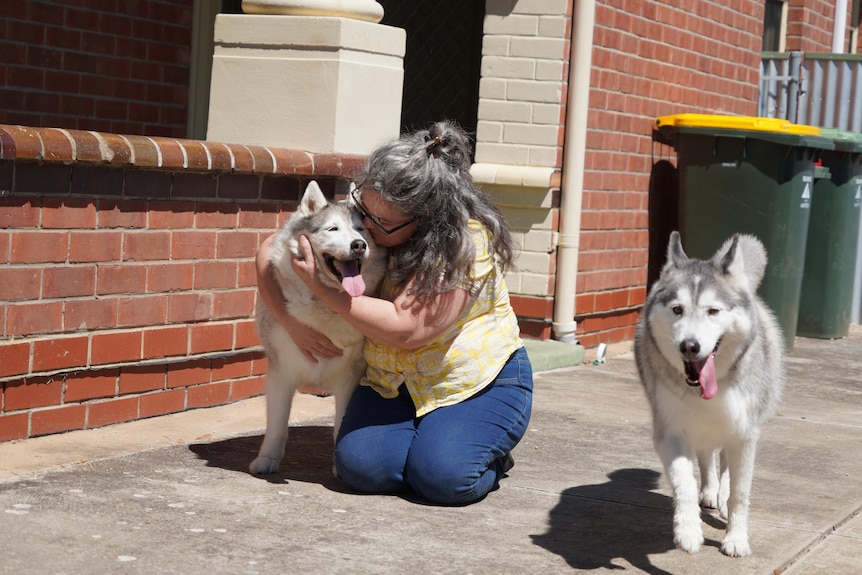 A woman with long grey curly hair kneels on the ground and cuddles her husky