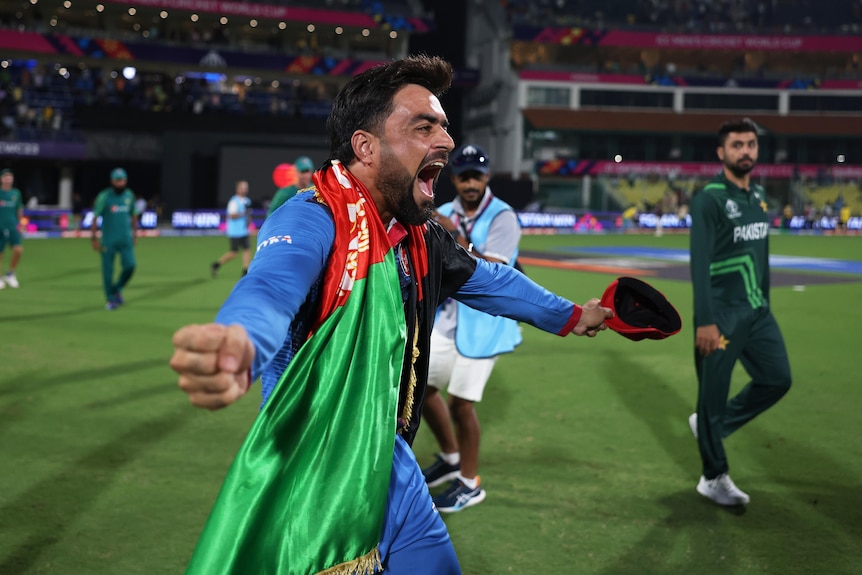 Rashid Khan yells in delight with teammates with an Afghanistan flag draped over him