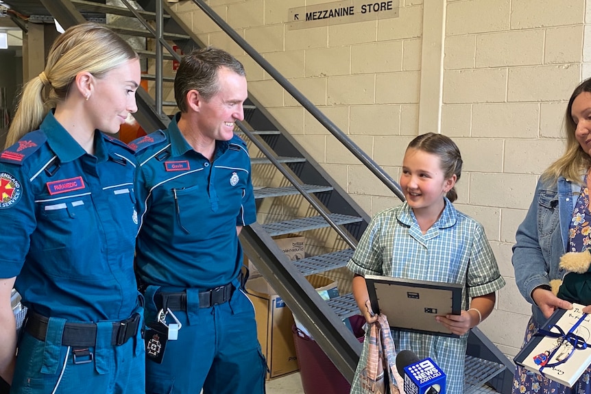 Two paramedics smile at a young girl holding a certificate of appreciation