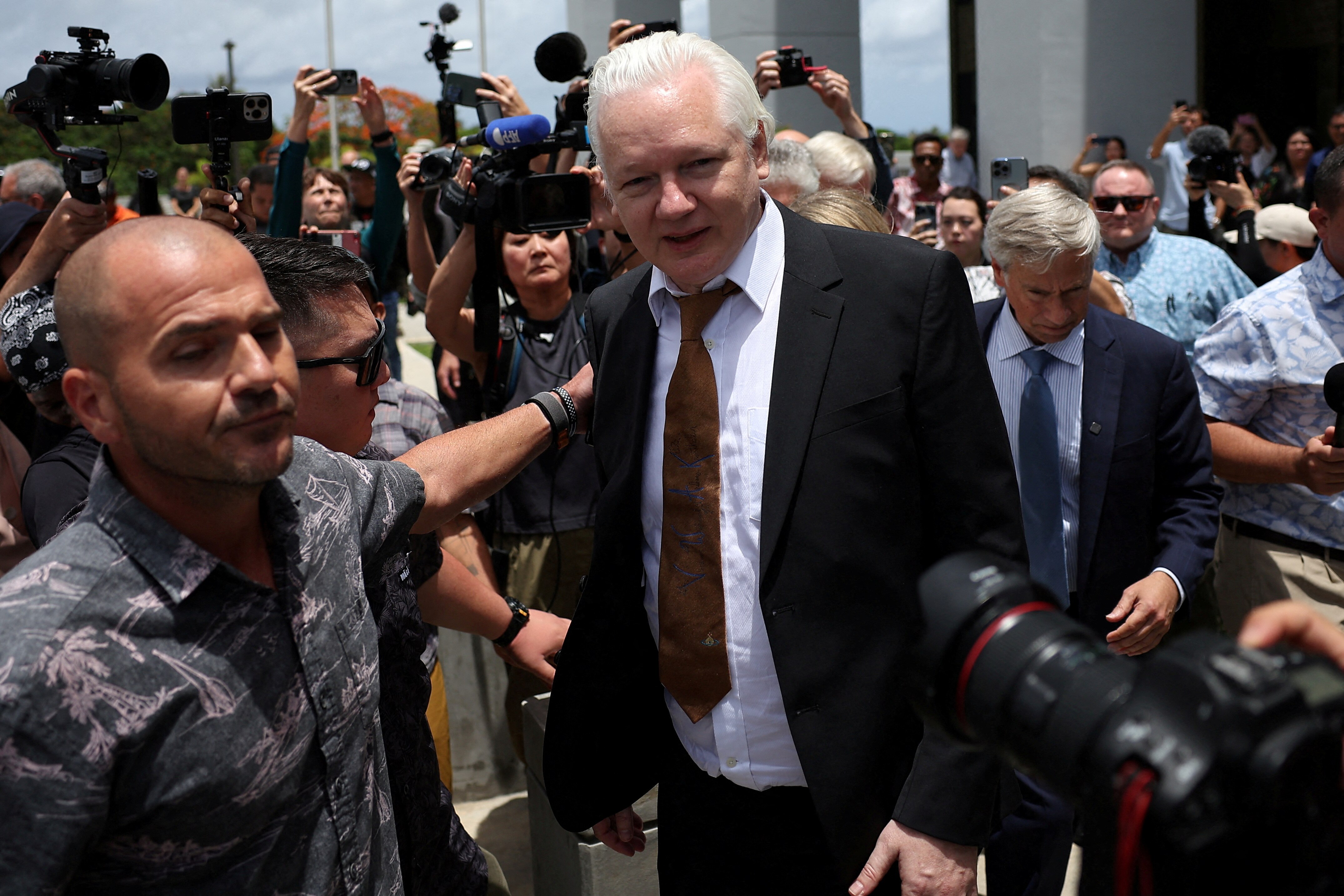 A middle-aged white man with white hair in a suit walks out of court surrounded by reporters.