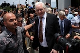 A middle-aged white man with white hair in a suit walks out of court surrounded by reporters.