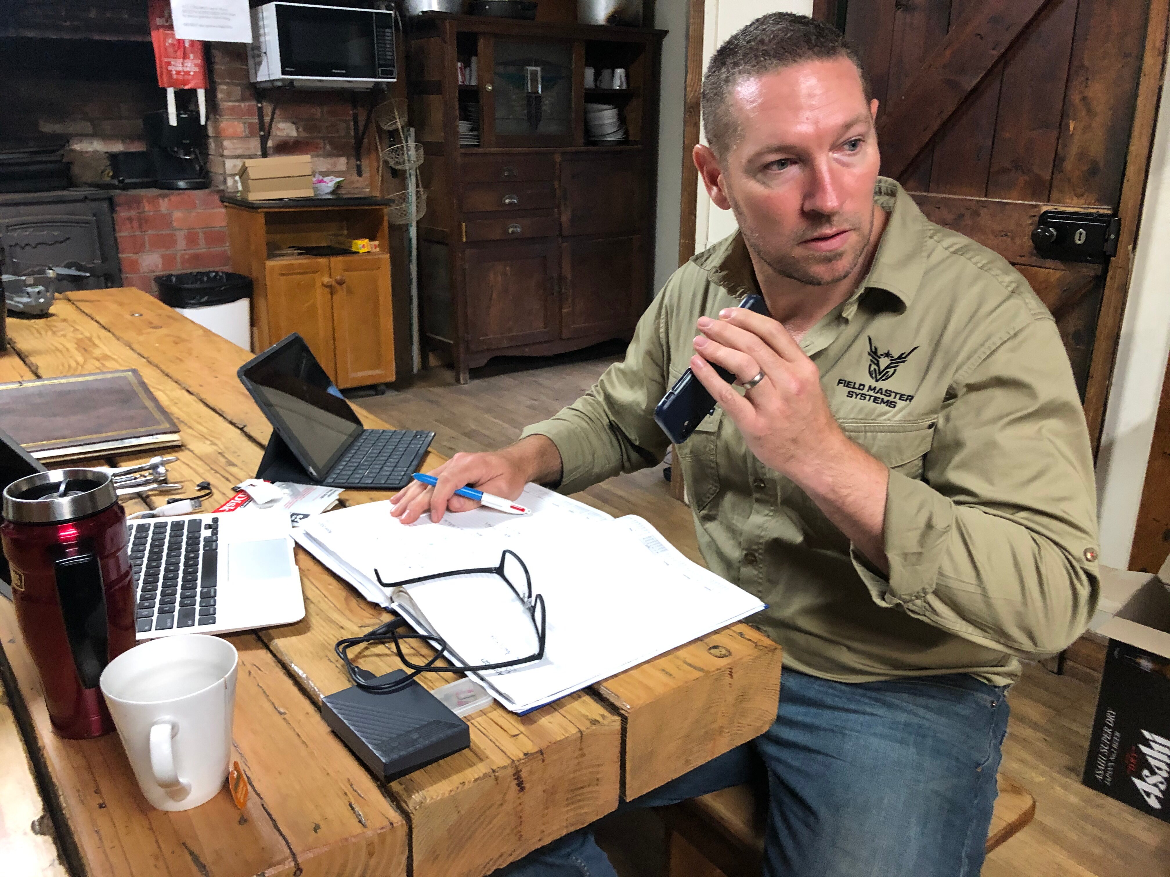A man in a khaki shirt holds a phone, sitting at a wooden table with laptops, papers and coffee cups in front of him.