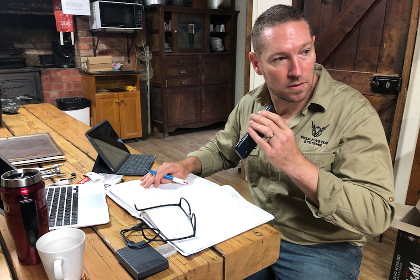 A man in a khaki shirt holds a phone, sitting at a wooden table with laptops, papers and coffee cups in front of him.