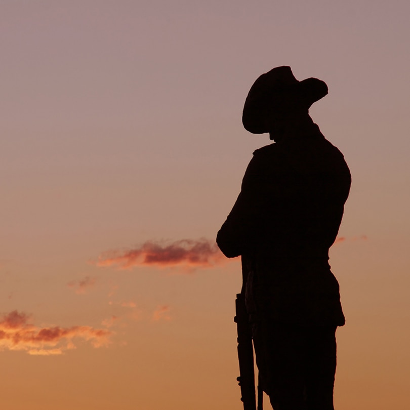 A soldier stands in silhouette, head bowed with a clear dawn sky in the background 