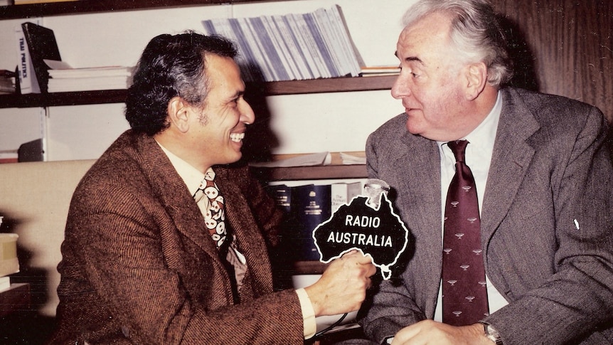 Man holding microphone with Radio Australia logo interviews Whitlam in an office.