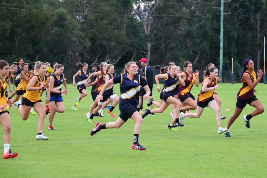 More than 20 girls in their Aussie Rules training gear race across an oval.