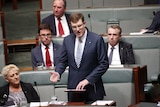 A bespectacled and besuited man with short, ginger hair stands in parliament, speaking.