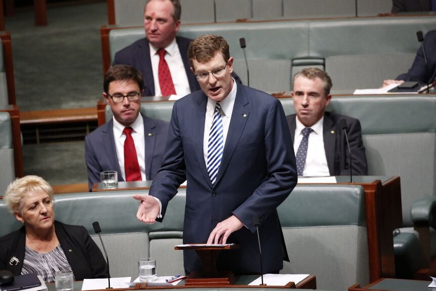 A dark-haired man in a dark suit stands to speak in parliament.