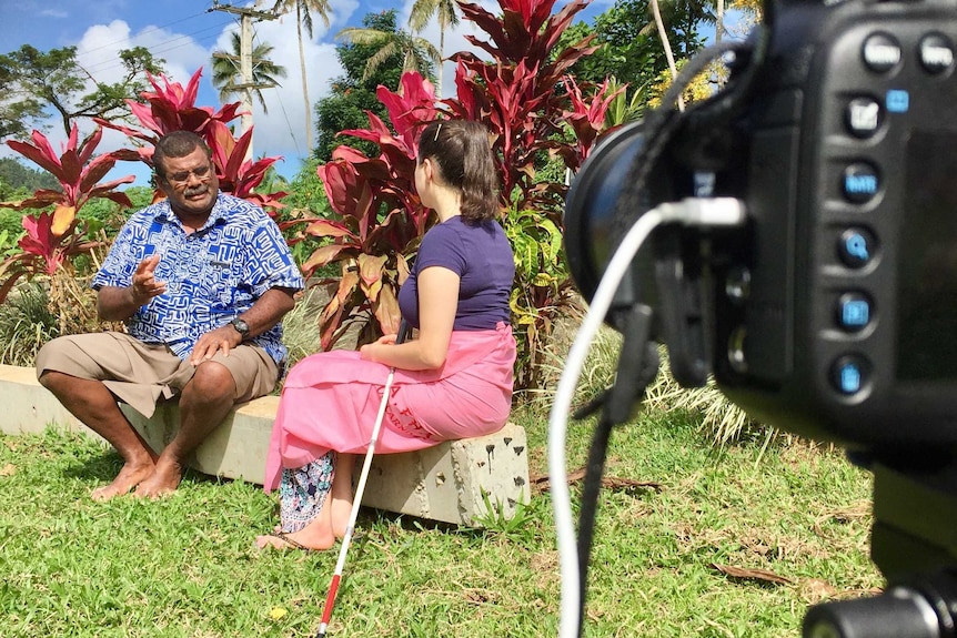 Tight shot of camera filming Nastasia Campanella from behind doing an interview with a Fijian man with her cane at her side.