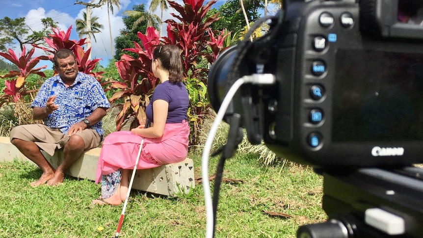Tight shot of camera filming Nastasia Campanella from behind doing an interview with a Fijian man with her cane at her side.