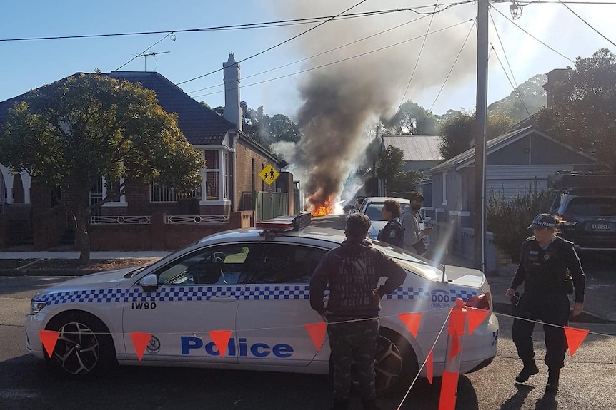 People watch on as flames and smoke billow from a burning car in a narrow laneway