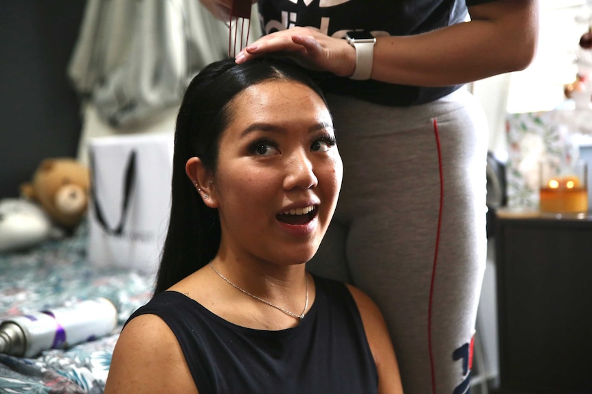 A woman sits and smiles in a chair, while another woman touches her hair