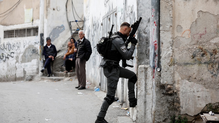 A man in a dark green uniform carries a gun while walking along a street. 