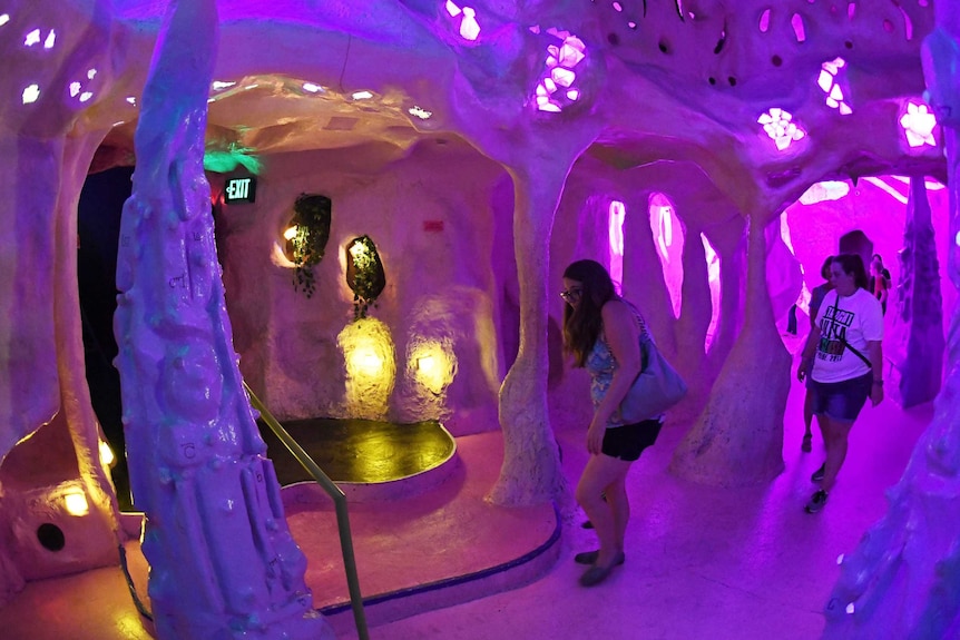 In a dark, purple-lit, cave-like room, people walk around fake stalactites.