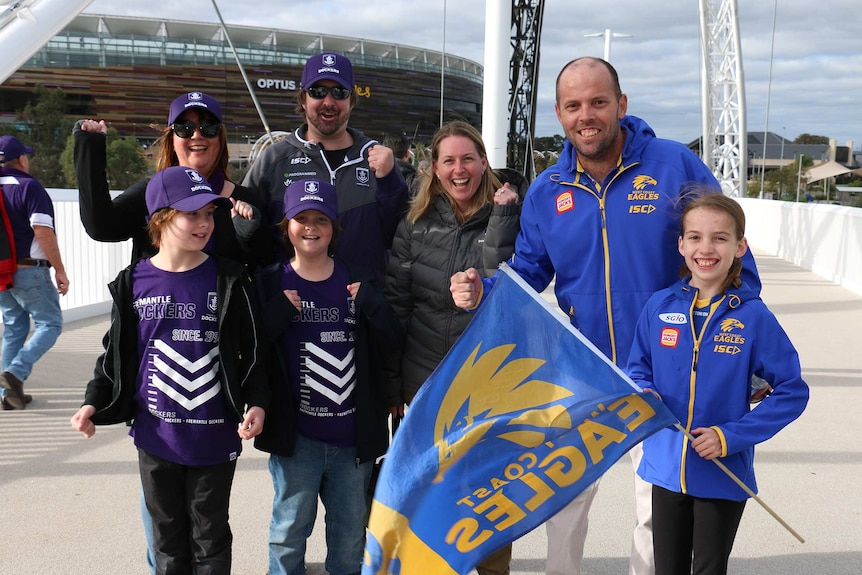 Two families, one clad in Dockers colours and the other in Eagles colours, stand on Matagarup bridge side by side cheering.