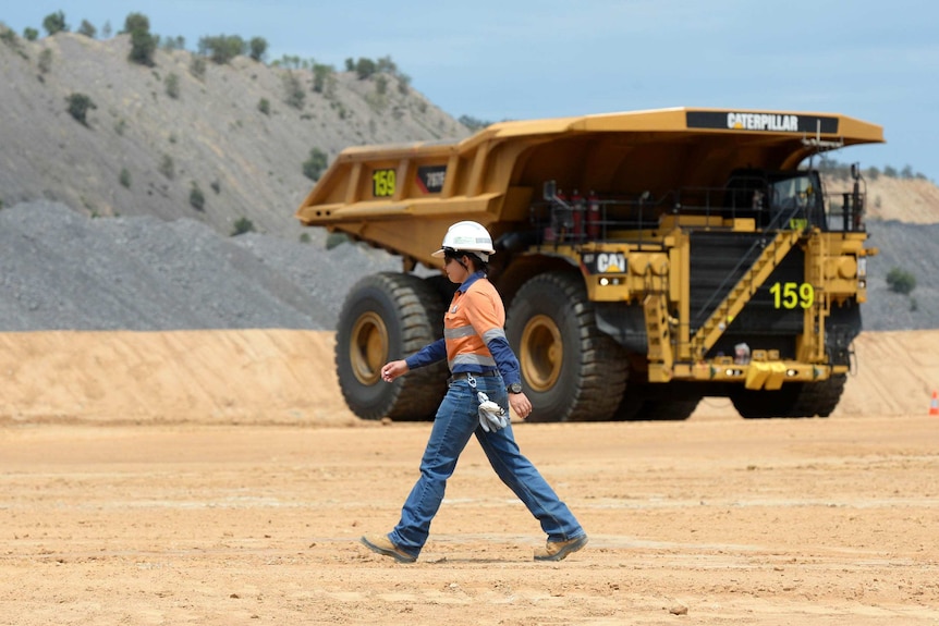 A woman in work clothes and a hard hat walks past a big mining truck.