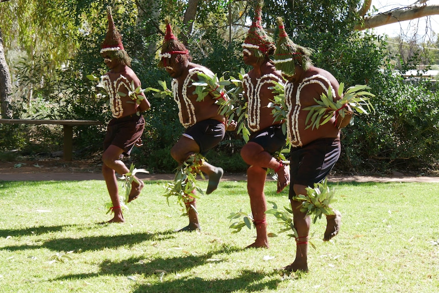 Four Indigenous men dance with their arms behind their backs, wearing leaves on their arms, legs, and heads.